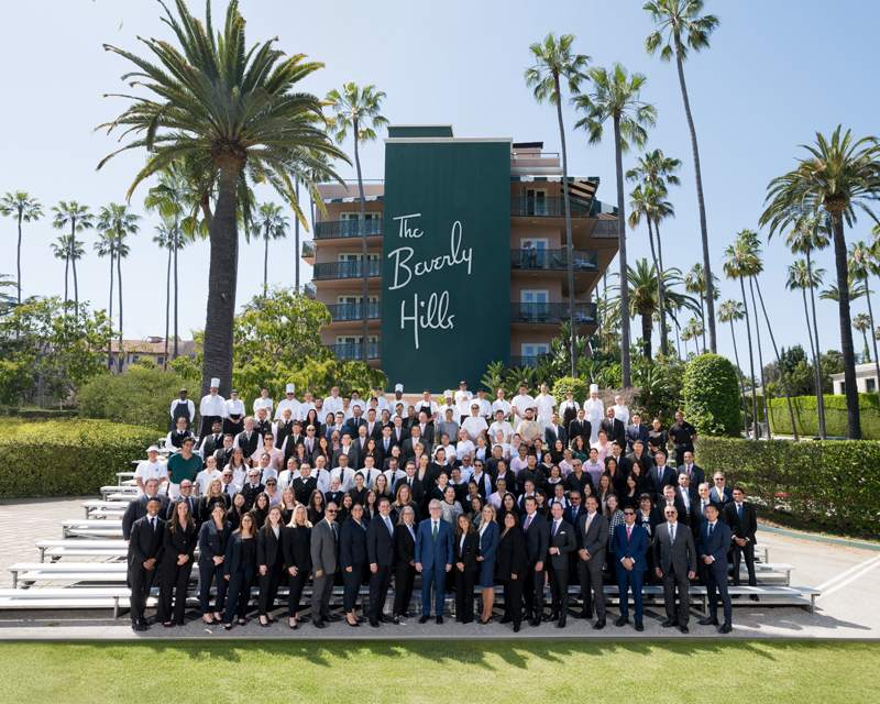 A large hotel team in dark suits and chef whites posed on outdoor bleachers under palm trees.