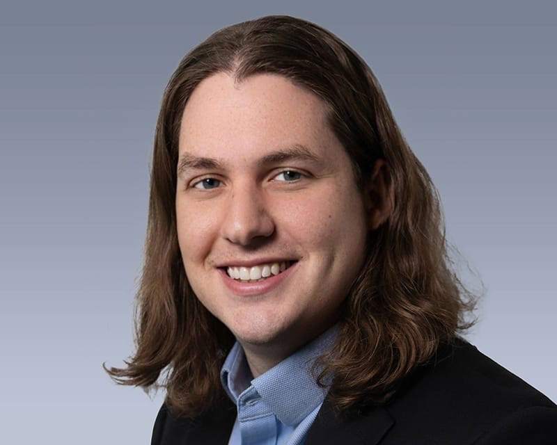 A long-haired man in a dark jacket and blue shirt smiling warmly against a grey studio background.