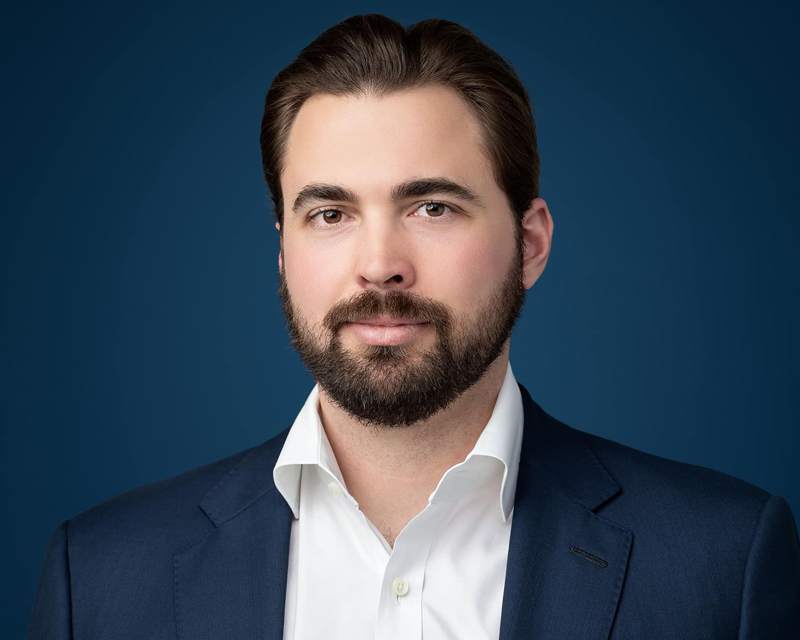 The same man in a navy suit and open white shirt against a deep blue studio background.