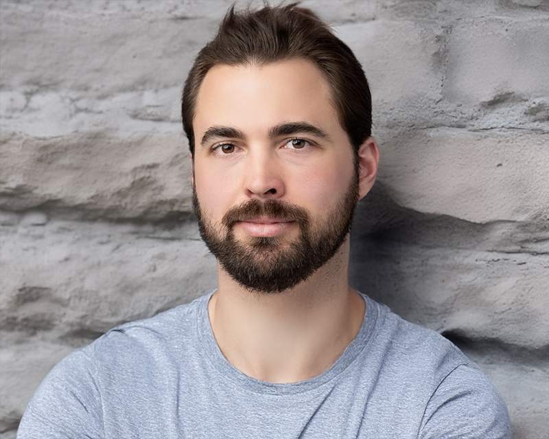 A bearded man in a grey T-shirt photographed in front of a pale stone wall.